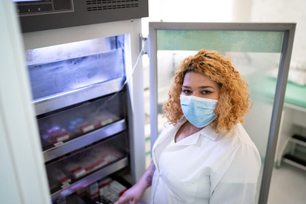 A female clinician stands inside the open door of a vaccine storage unit equipped with a data logger.