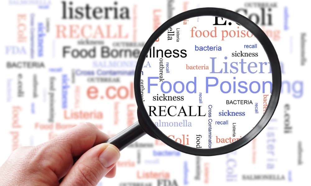 In the background, words such as foodborne illness, recall, food poisoning, and others appear on a white background in black, red, and blue. A hand holds a magnifying glass, enlarging some of the words, including bacteria, sickness, and recall.